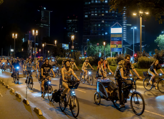 Gruppe von Radfahrenden bei Nacht auf einer Straße in Frankfurt. Die Fahrräder sind mit blauem und weißem Licht beleuchtet. Im Hintergrund sind Hochhäuser und Straßenlaternen zu sehen. Die Teilnehmenden fahren verschiedene Fahrradtypen wie Lastenräder, Stadträder und normale Fahrräder.